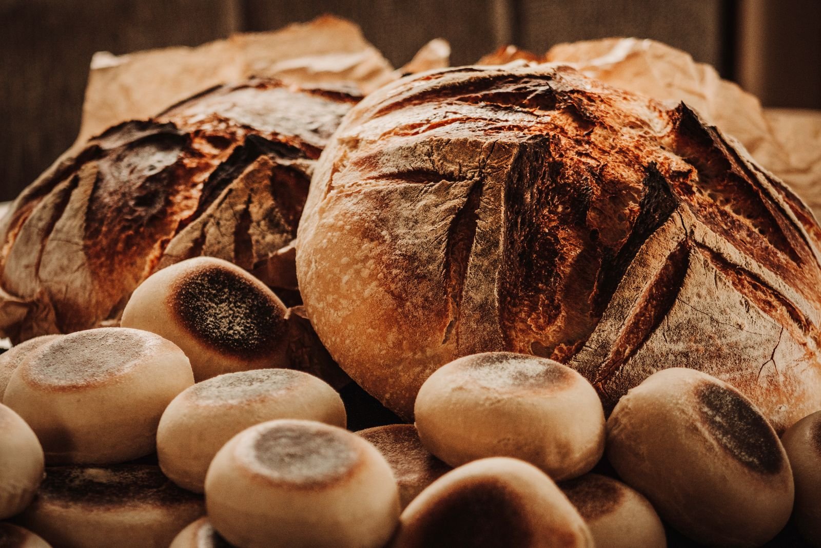 Rustic scored sourdough boules and flour-dusted buns piled on parchment in warm light